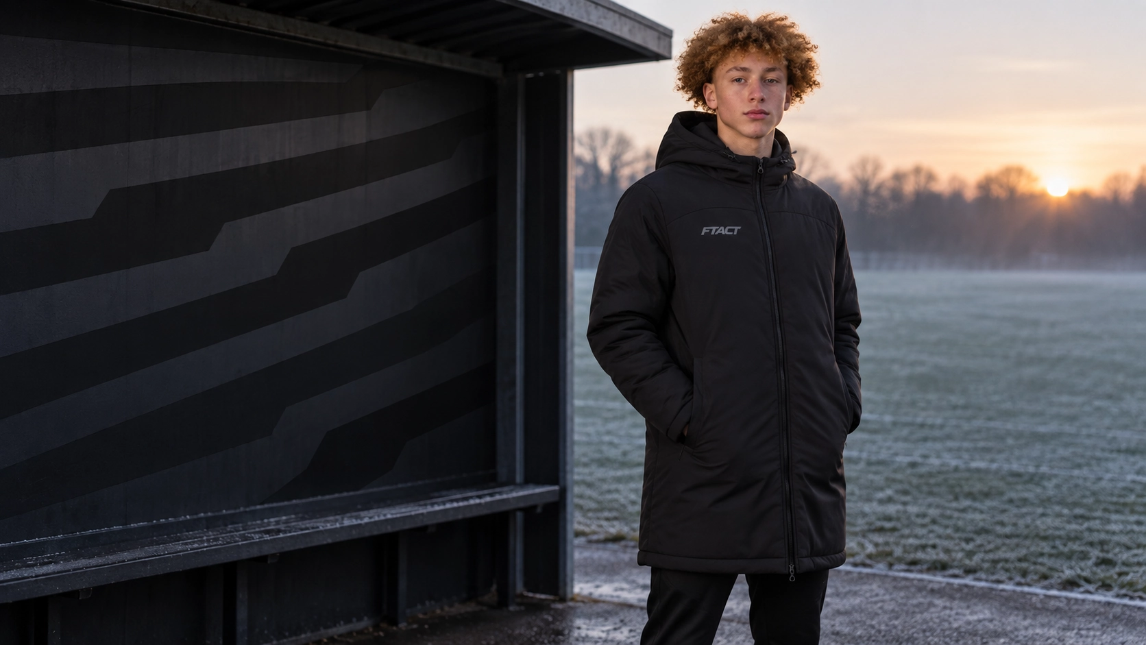 Athlete in a thigh-length black FTACT bench coat beside an empty football touchline shelter