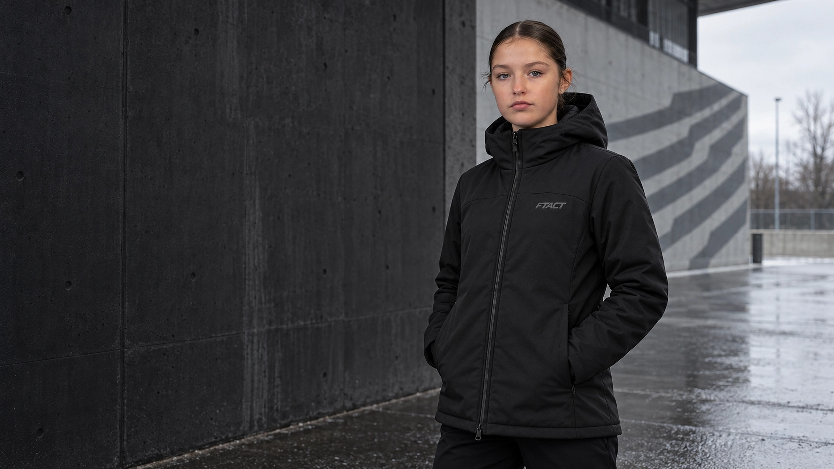 Athlete in a black FTACT winter parka at a rain-wet football facility entrance