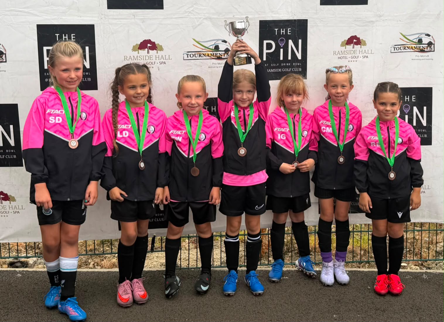 Young players celebrating with medals and a trophy in their pink FTACT training wear
